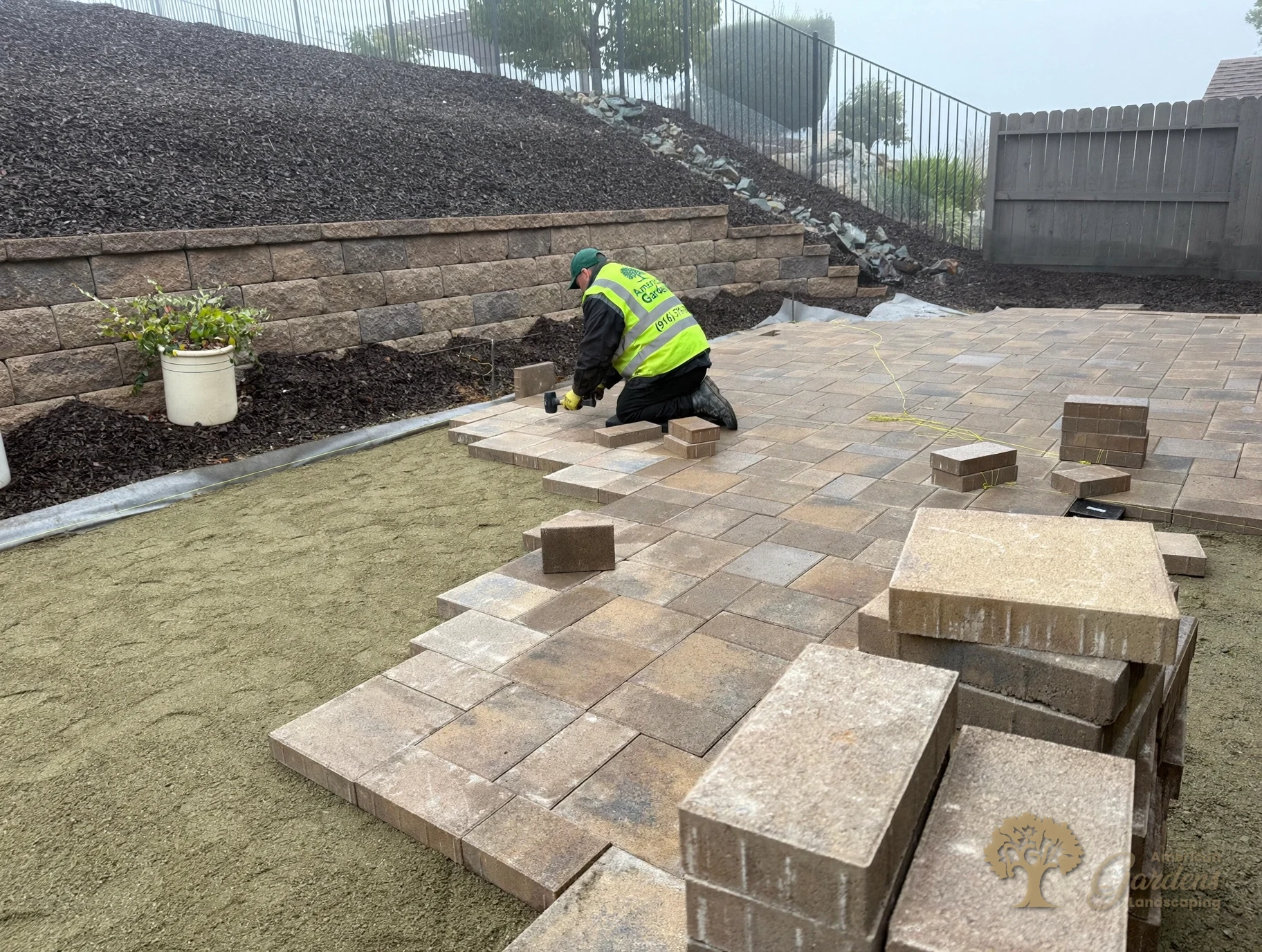Crew member setting pavers at the base of the capped block retaining wall that holds back the bark-covered slope in El Dorado Hills, by American Gardens Landscaping.