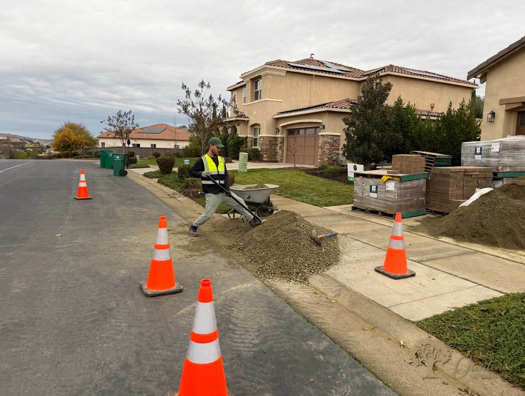 American Gardens Landscaping crew member shoveling delivered base rock into a wheelbarrow between safety cones, with pallets of pavers ready, in El Dorado Hills.