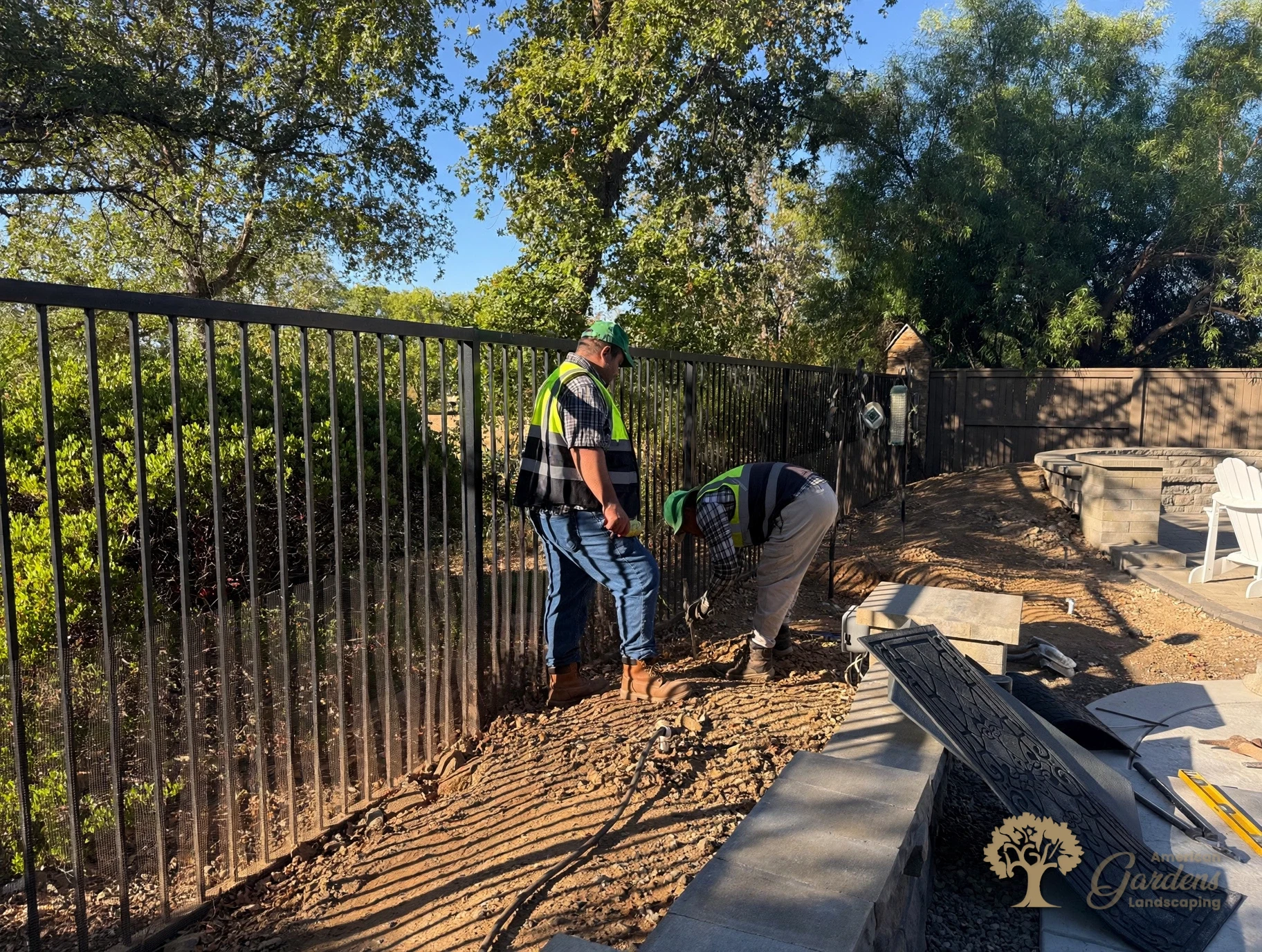 Two crew members in branded vests grading and fitting along a black metal fence in a Serrano backyard in El Dorado Hills, by American Gardens Landscaping.
