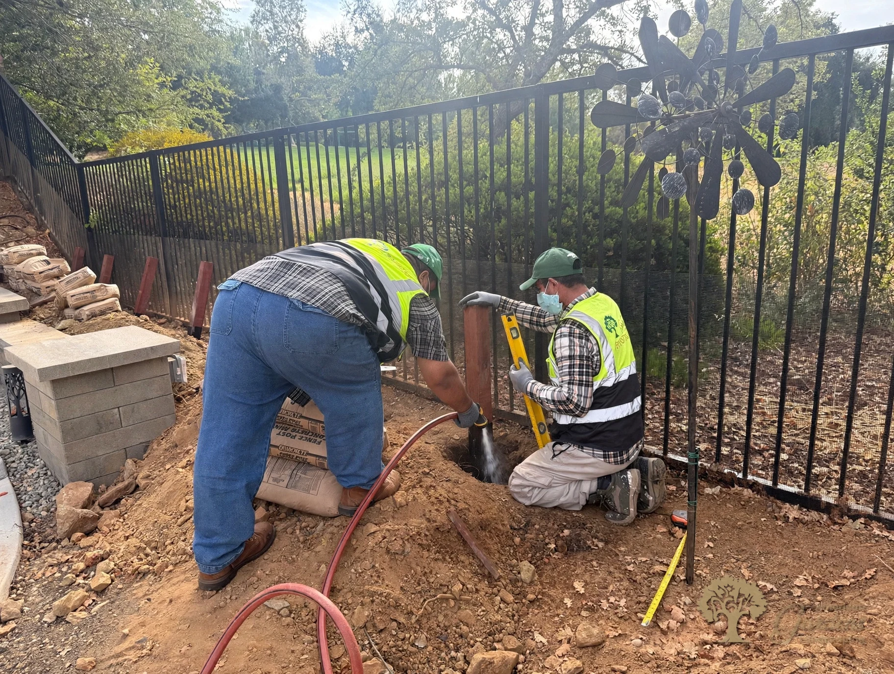 Crew wetting down concrete around a leveled fence post beside a black metal fence in Serrano, El Dorado Hills, by American Gardens Landscaping.