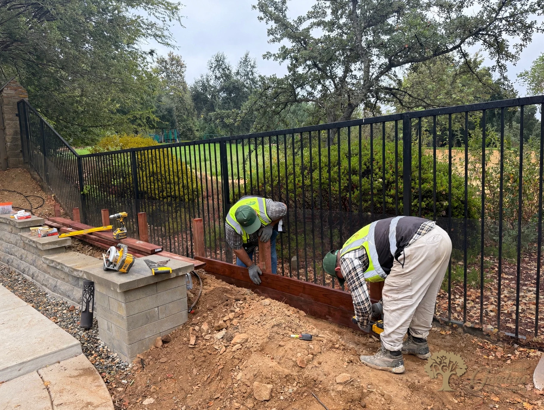 Wide view of the crew installing a stained board along new posts below a black metal fence in a Serrano backyard in El Dorado Hills, by American Gardens Landscaping.