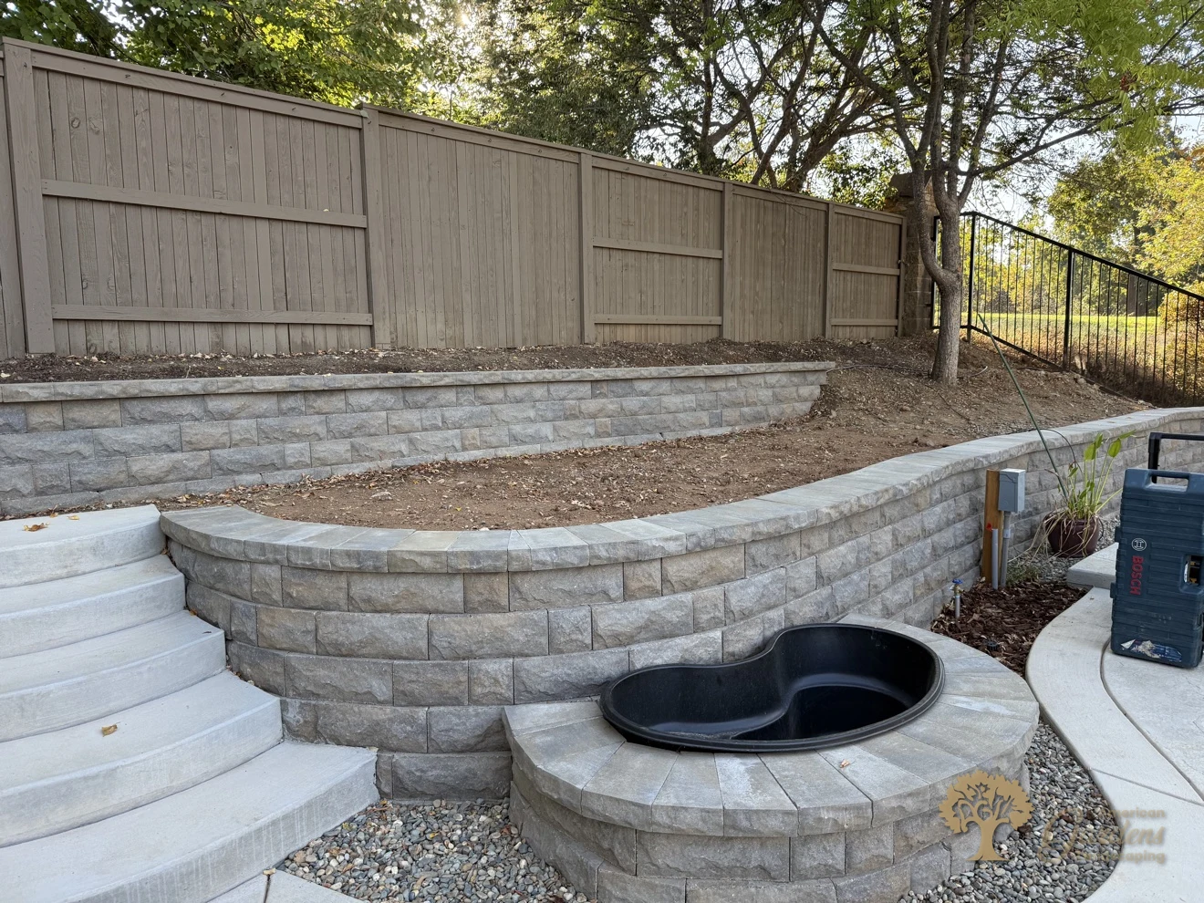 Curved block retaining walls, concrete steps and a pond basin surround viewed from the steps side in a Serrano backyard in El Dorado Hills, by American Gardens Landscaping.