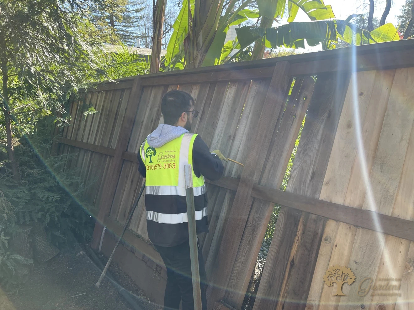American Gardens crew member in a branded hi-vis vest repairing a wooden fence in Fair Oaks