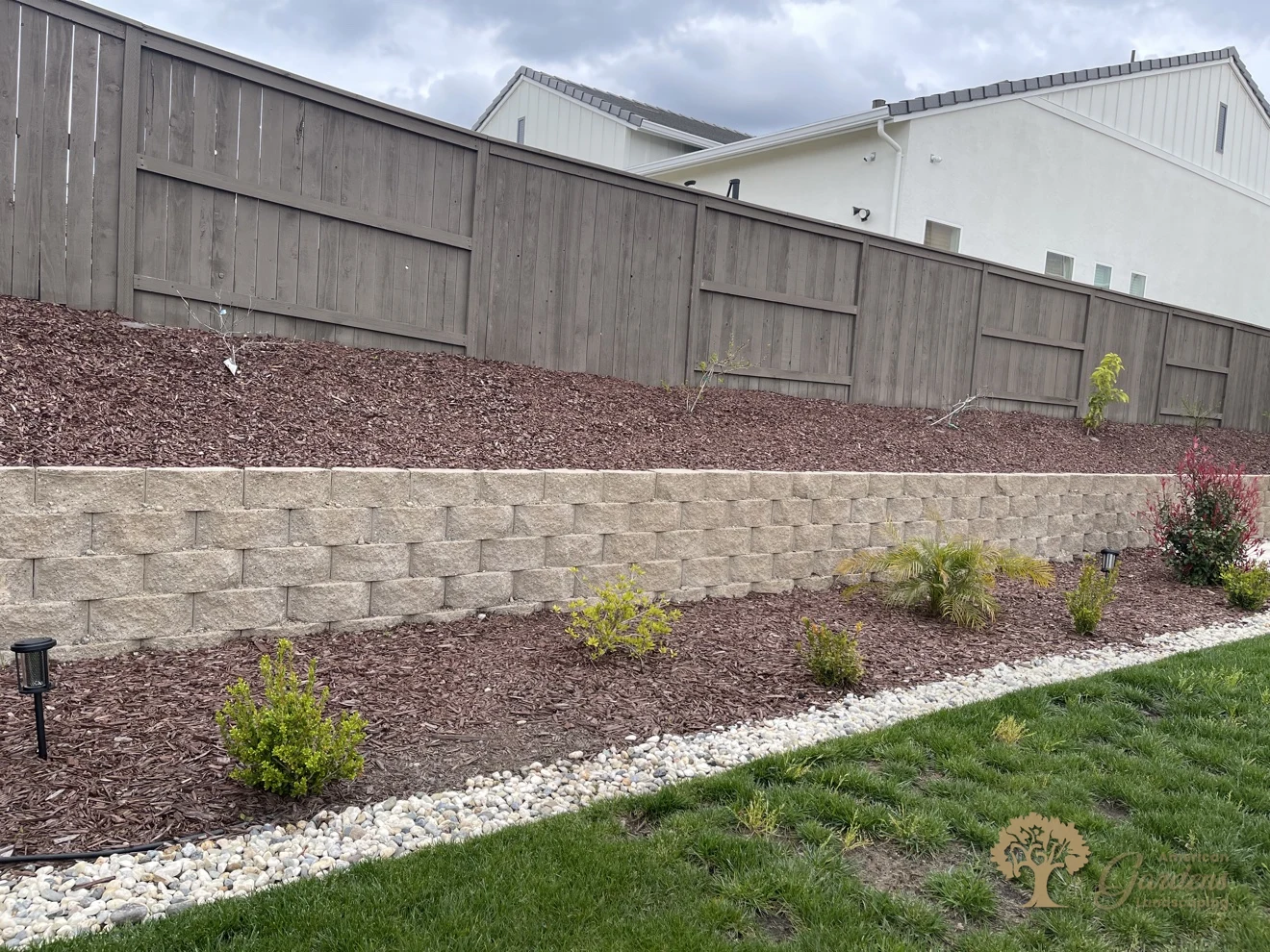 Long concrete block retaining wall holding a mulched slope with new plantings and a river rock border in Rocklin