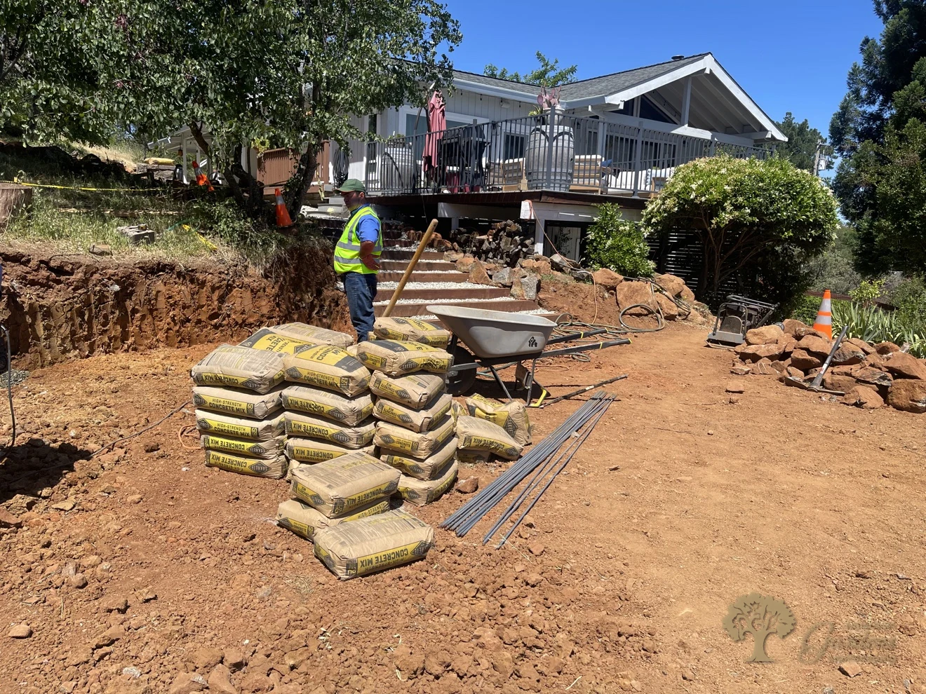 Crew member standing by concrete mix bags and rebar staged for a segmental block retaining wall build below a house deck in Shingle Springs
