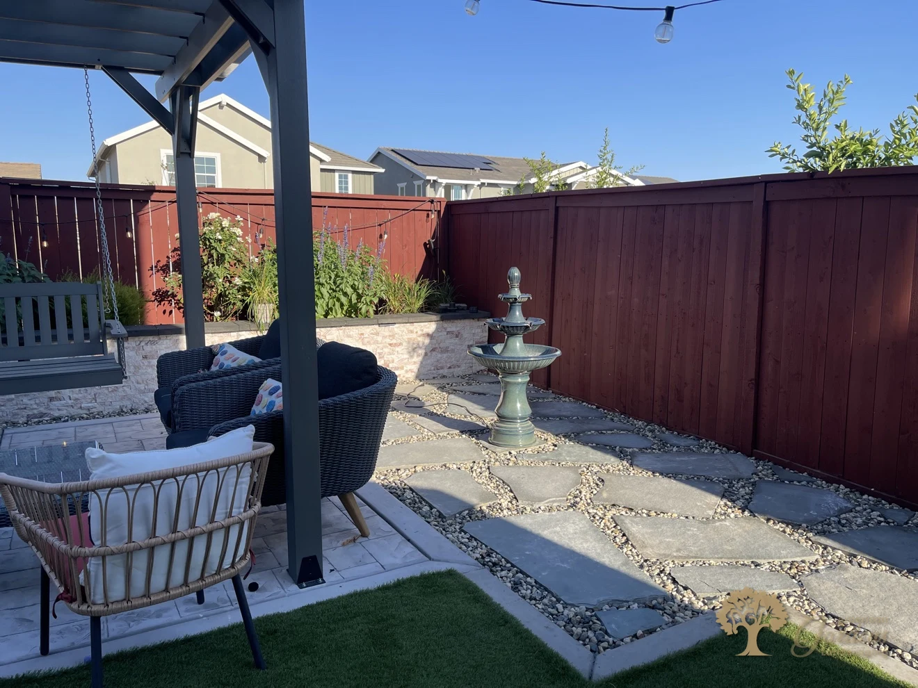 Under the pergola in a Lincoln backyard, a hanging swing and wicker seating overlook the bluestone patio and tiered fountain by the red-stained fence, by American Gardens Landscaping