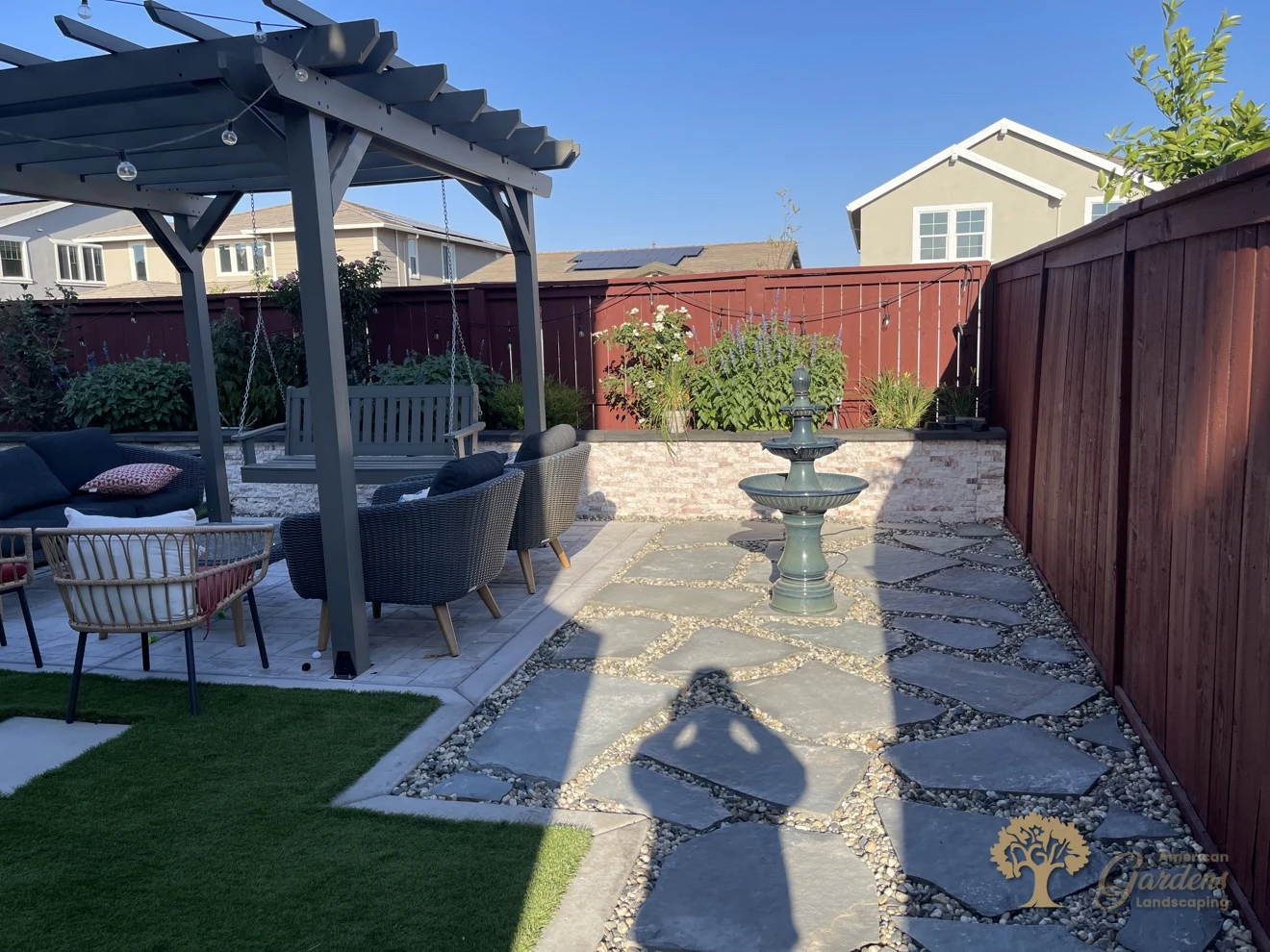 A tiered fountain on the bluestone patio in a Lincoln backyard, with a pergola, swing and seating to the side and artificial turf in front, by American Gardens Landscaping