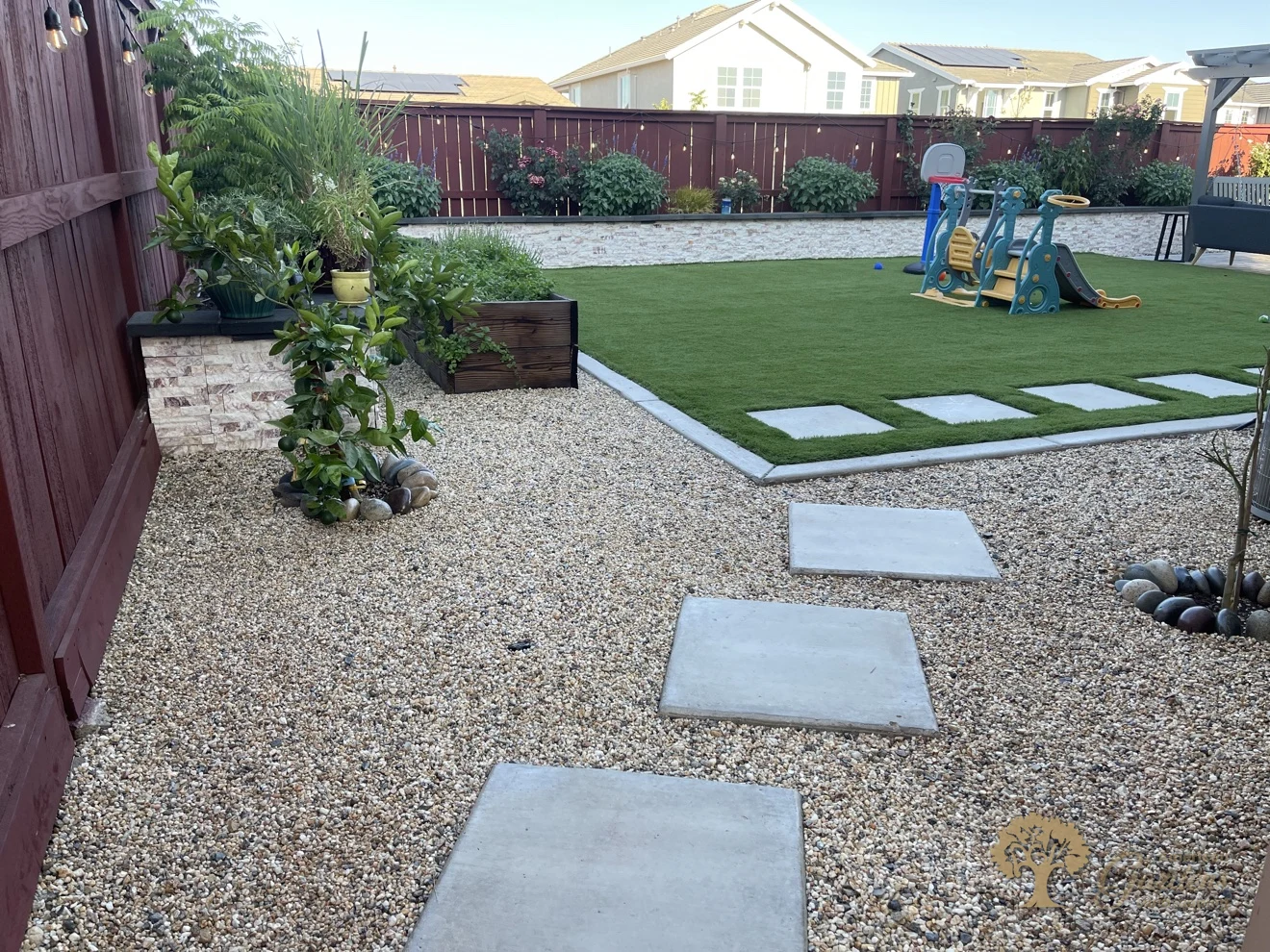 The artificial turf lawn in a Lincoln backyard with square concrete stepping stones crossing the gravel side area beside the red-stained fence, by American Gardens Landscaping