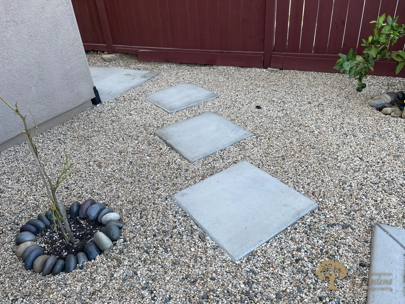A gravel side area with square concrete stepping stones and a small tree ringed with river rock beside the red-stained fence in a Lincoln backyard, by American Gardens Landscaping