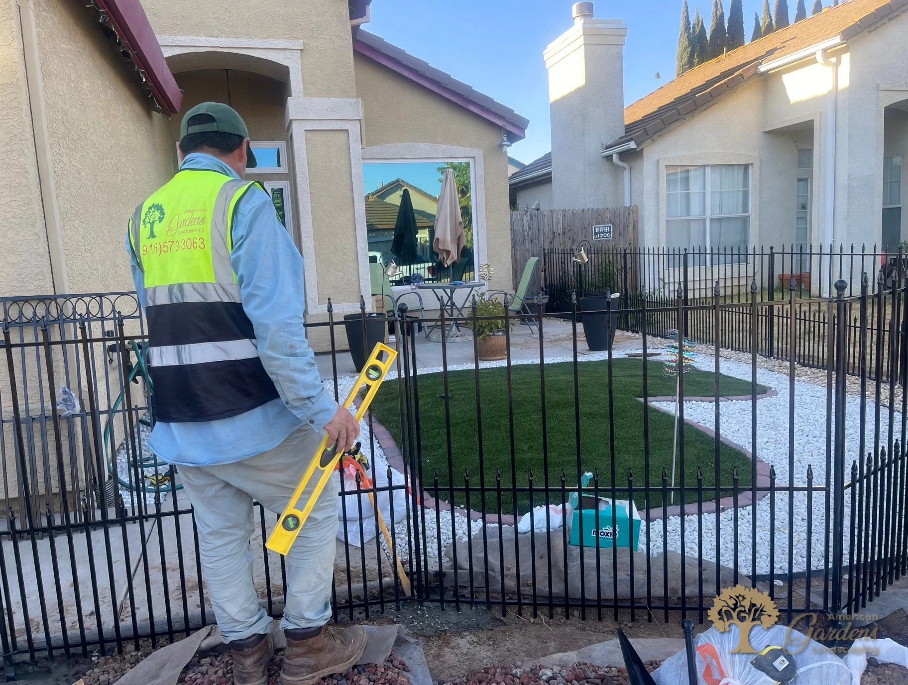 American Gardens Landscaping crew member in a company vest holding a yellow level beside a black metal fence in a front yard with white rock and a small green patch