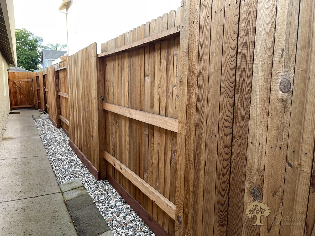 Stained vertical-board fence along a side yard with a gravel strip and concrete walkway, built by American Gardens Landscaping