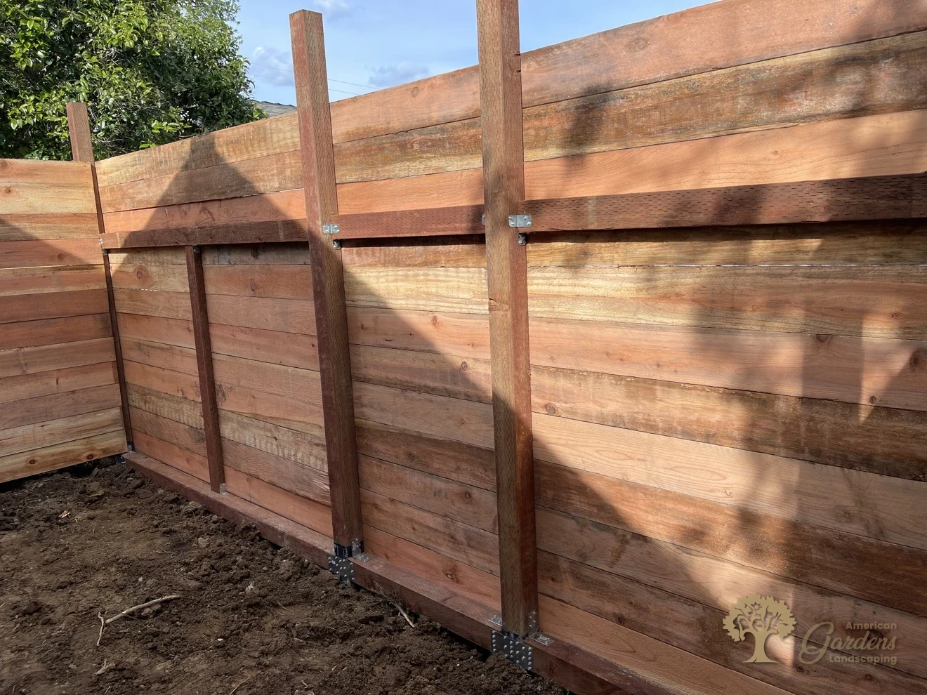 Back side of a horizontal redwood fence showing pressure-treated posts on black post-base brackets, rails and Simpson steel connectors, built by American Gardens Landscaping