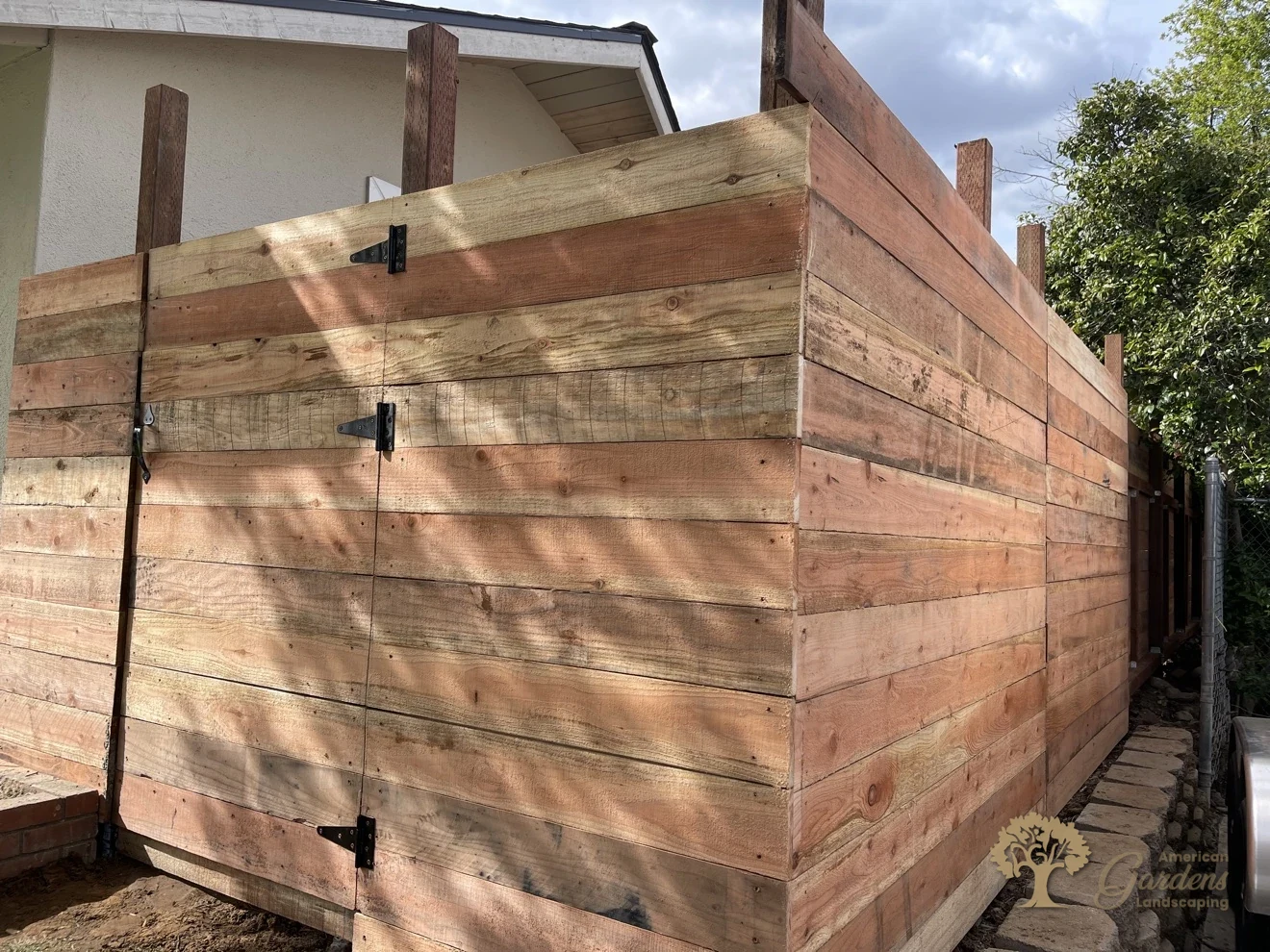 Double gate with black T-hinges on a horizontal redwood board fence at the corner of a house, built by American Gardens Landscaping