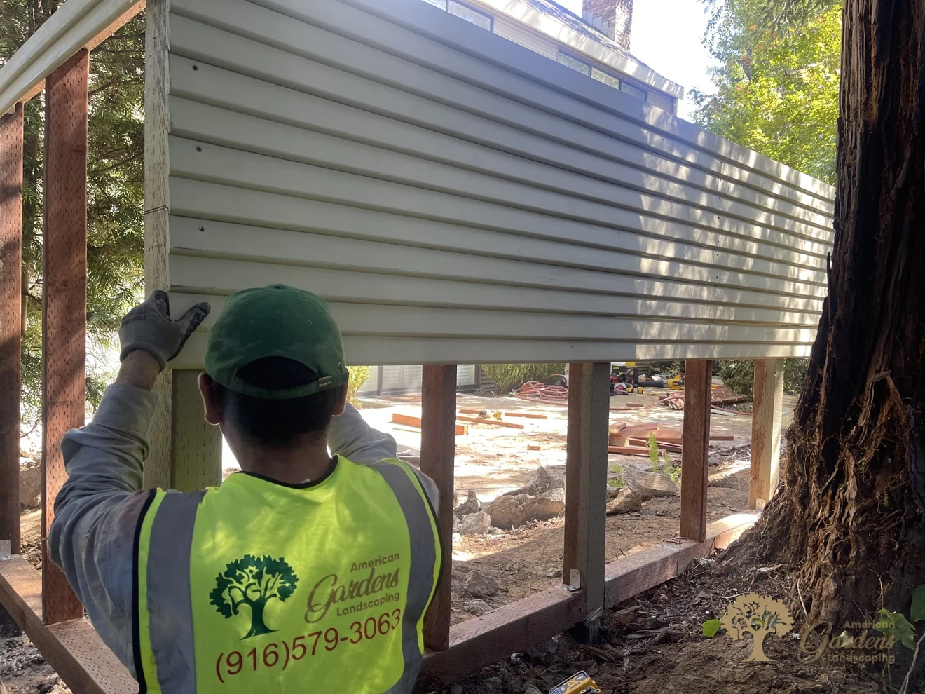 An American Gardens Landscaping crew member seen from behind in a company vest with the logo and phone number, holding a horizontal fence panel section into place between the posts in Sacramento