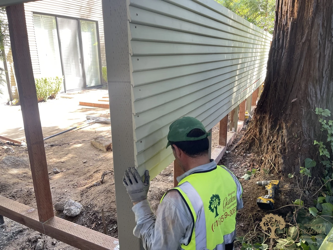 An American Gardens Landscaping crew member in a company vest and green cap fitting a horizontal fence panel section against a post beside a large redwood tree in Sacramento