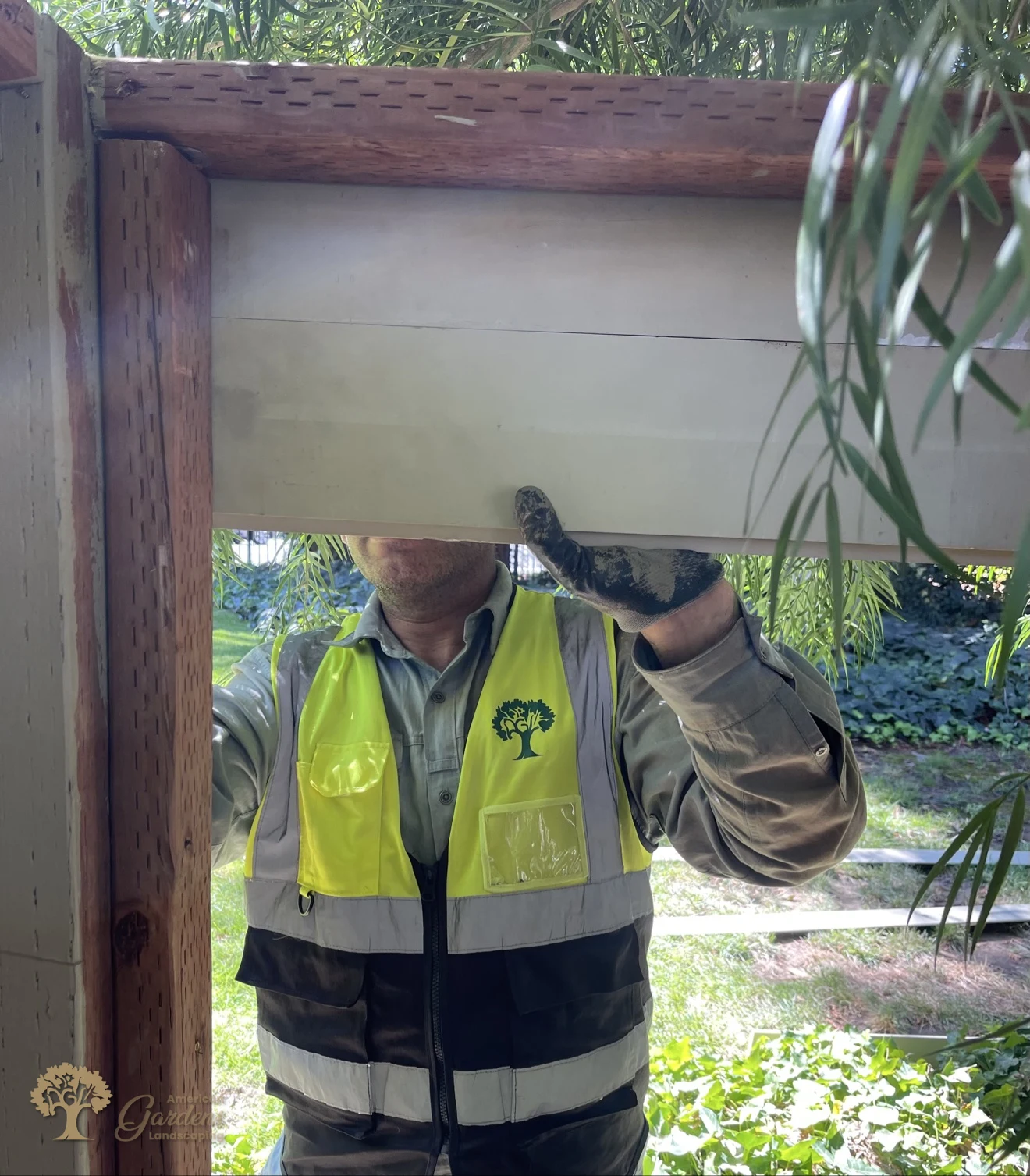 An American Gardens Landscaping crew member in a company vest holding a horizontal fence panel section up into the frame in Sacramento