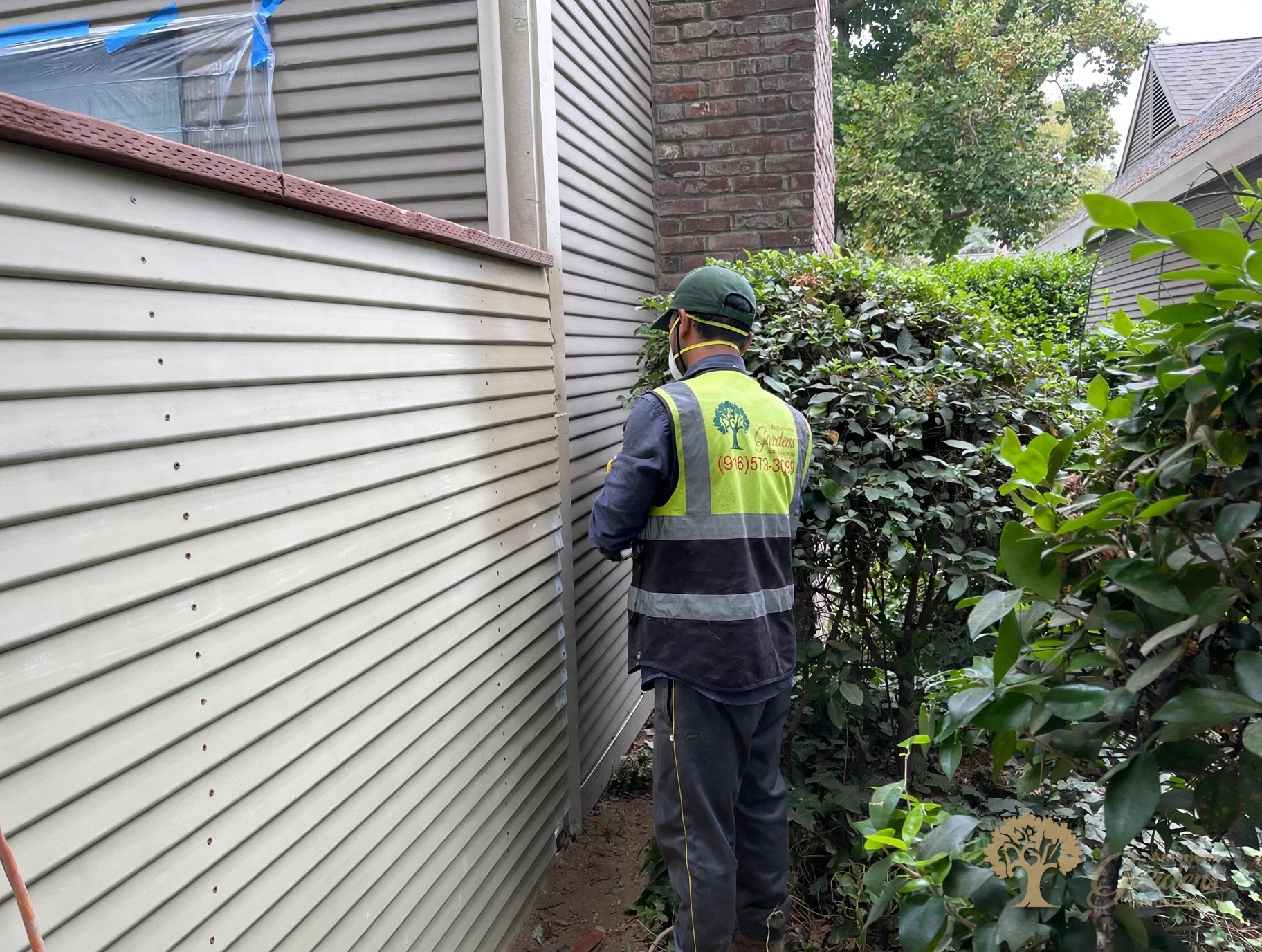 An American Gardens Landscaping crew member in a company vest beside the finished fence in Sacramento, with the windows masked in plastic and tape for painting