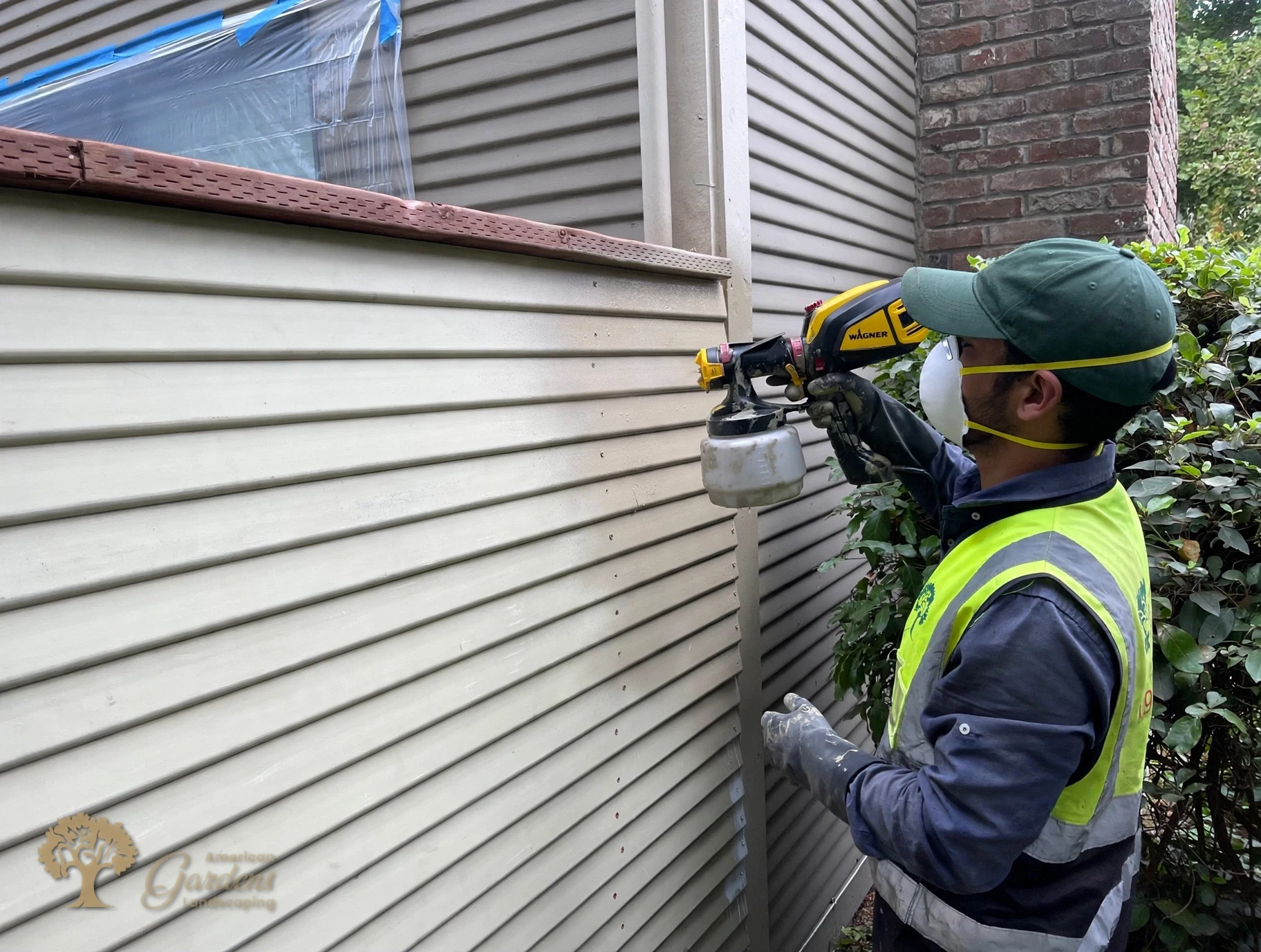 An American Gardens Landscaping crew member spray painting the new fence panels with a handheld sprayer in Sacramento, wearing a respirator mask