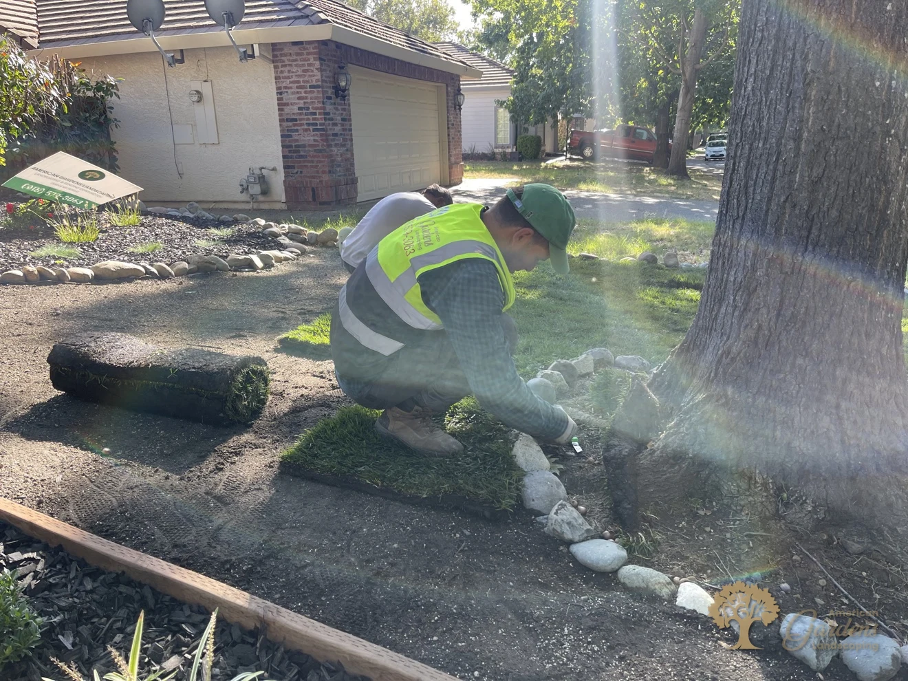 An American Gardens Landscaping crew member in a hi-vis vest kneeling to fit a piece of new sod at the oak tree's rock ring in a Folsom front yard