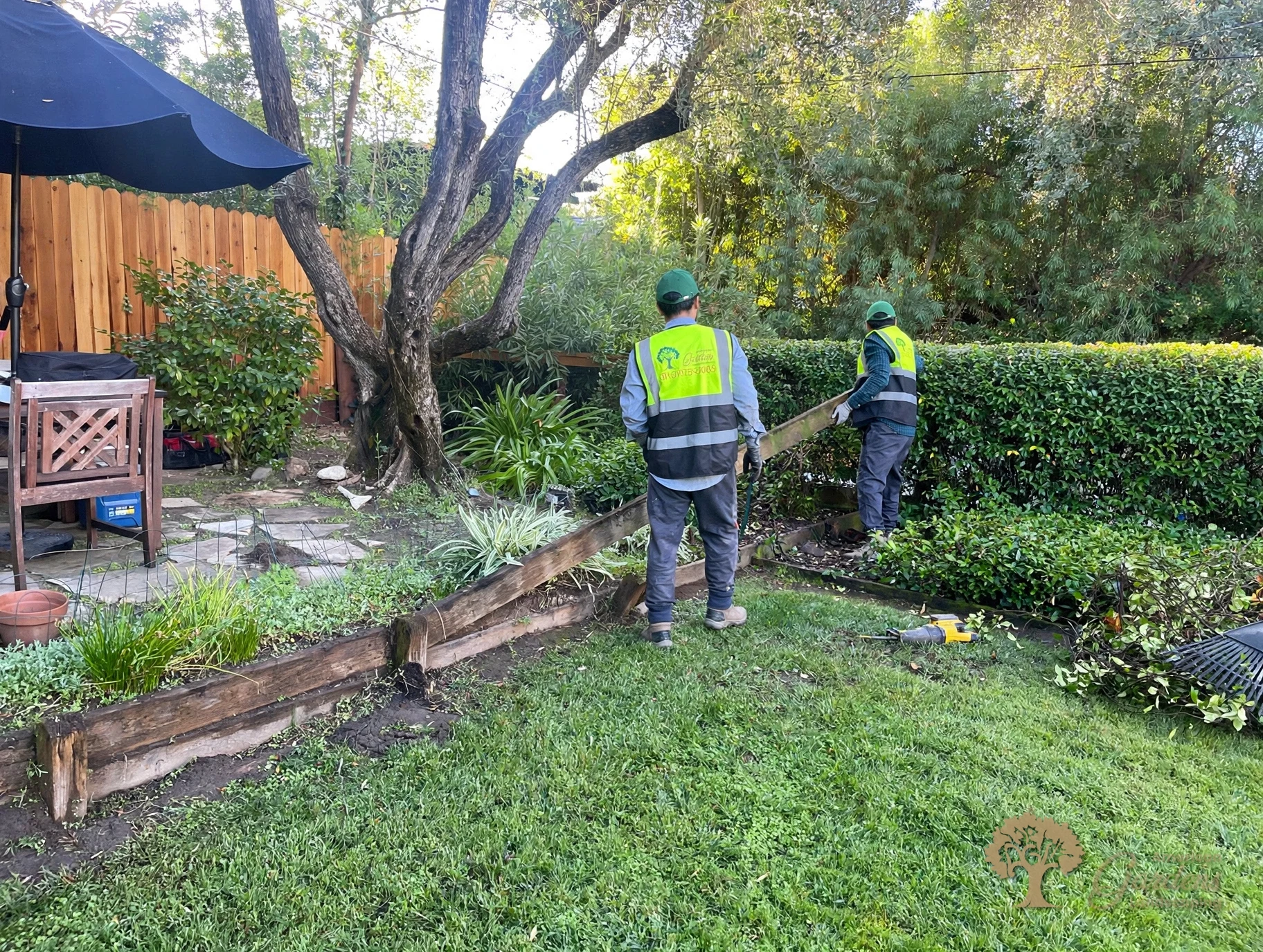Two American Gardens Landscaping crew members in company vests prying out the old railroad-tie timber edging at the start of a retaining wall replacement in Fair Oaks
