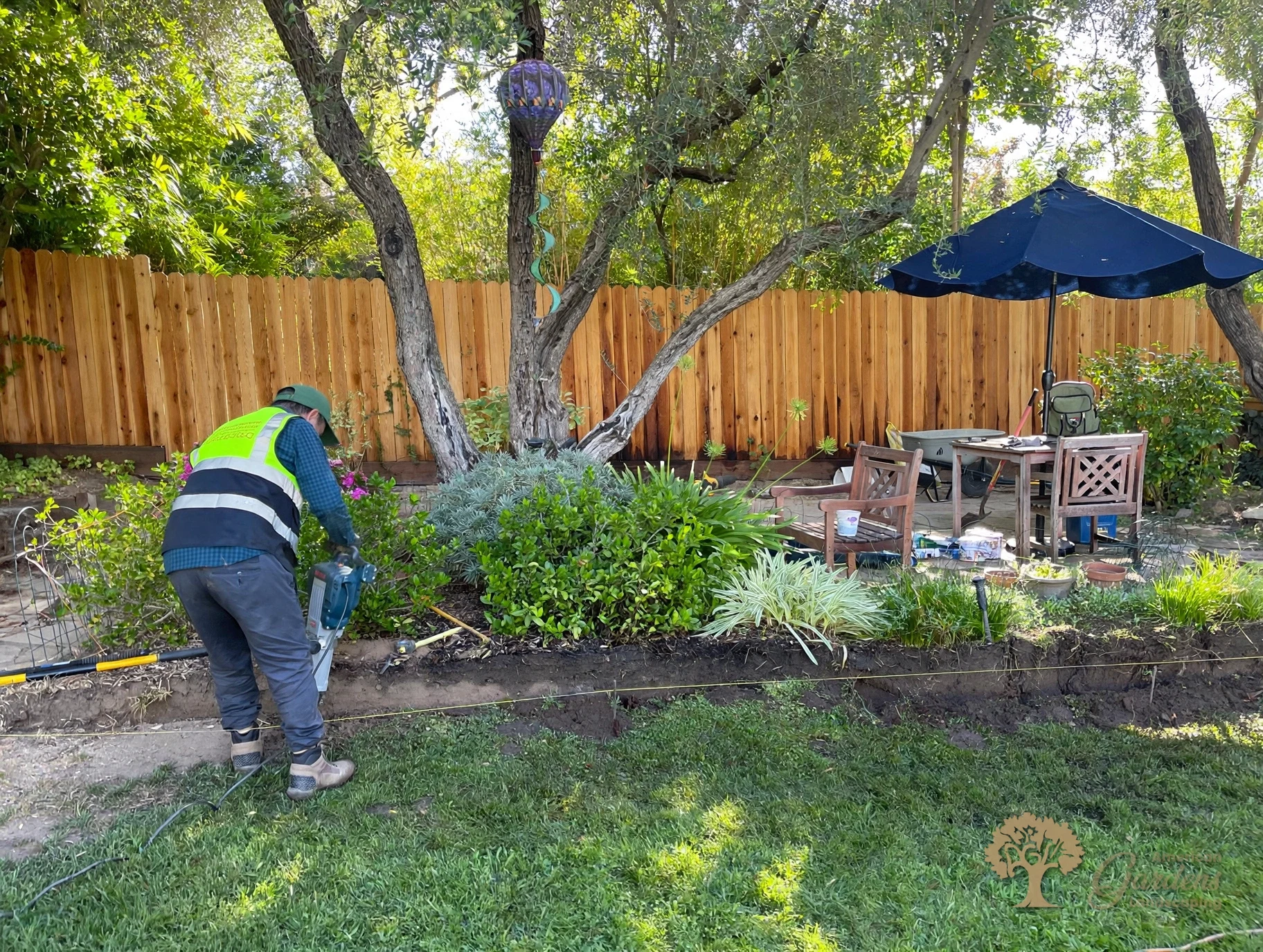An American Gardens Landscaping crew member in a company vest trenching with a jackhammer along the string line for the new retaining wall in Fair Oaks