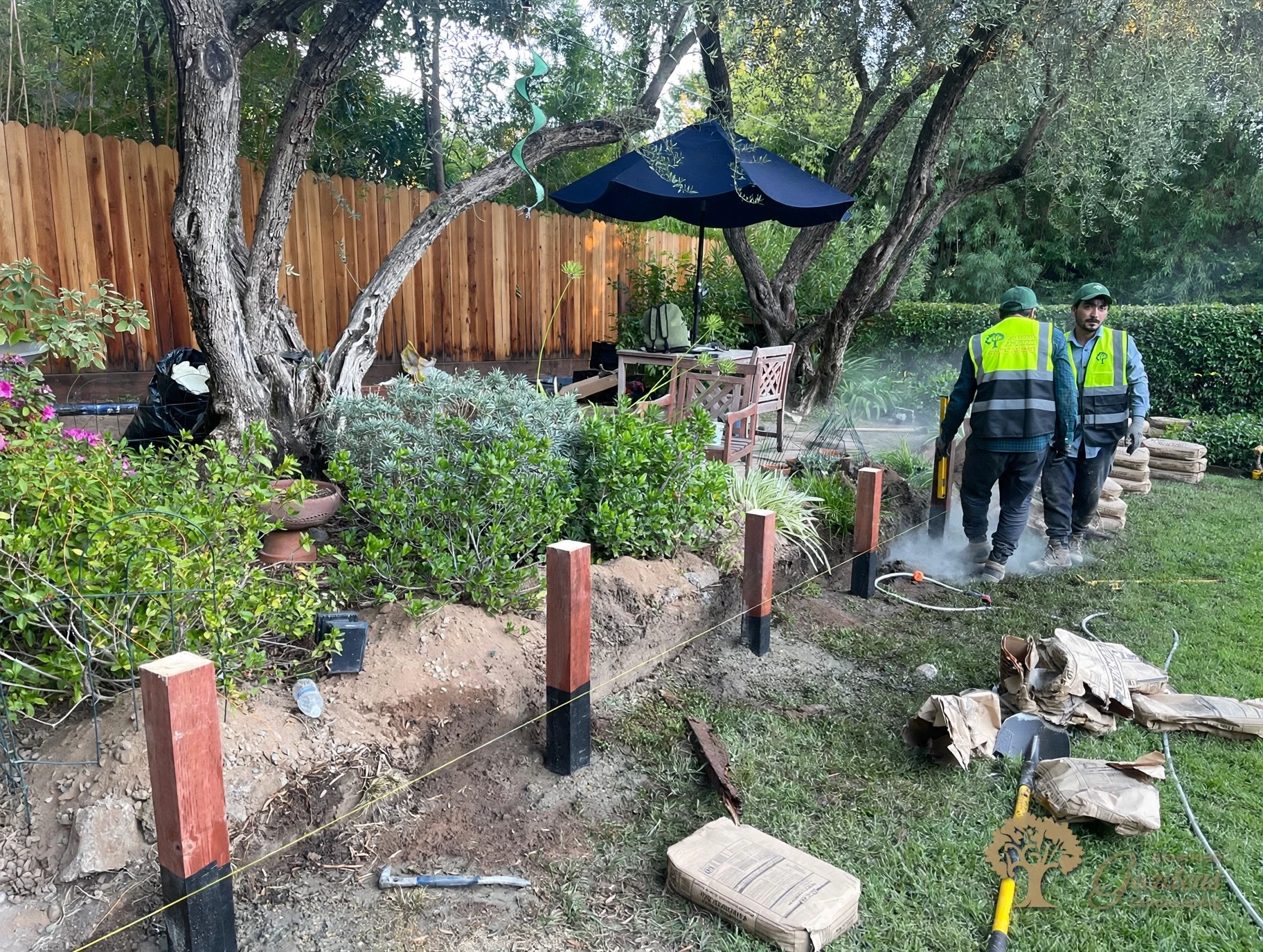 Two American Gardens Landscaping crew members plumbing a post with a yellow level, with the new posts set in a row on a string line with waterproof wrap on their bases and concrete bags at each hole in Fair Oaks