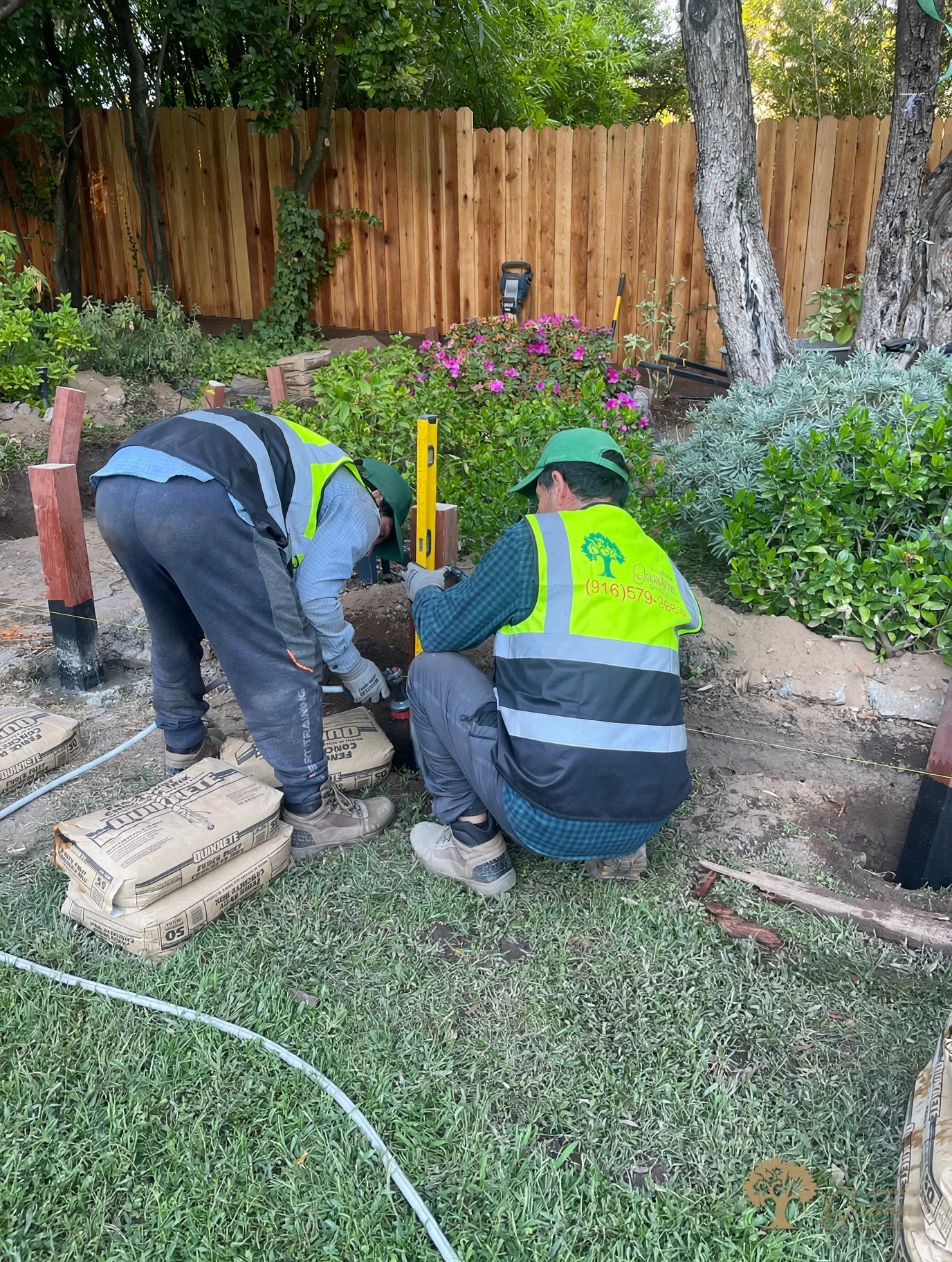 Two American Gardens Landscaping crew members setting a pressure-treated post with a yellow level in Fair Oaks, the post base wrapped in black waterproof wrap and concrete bags stacked beside it