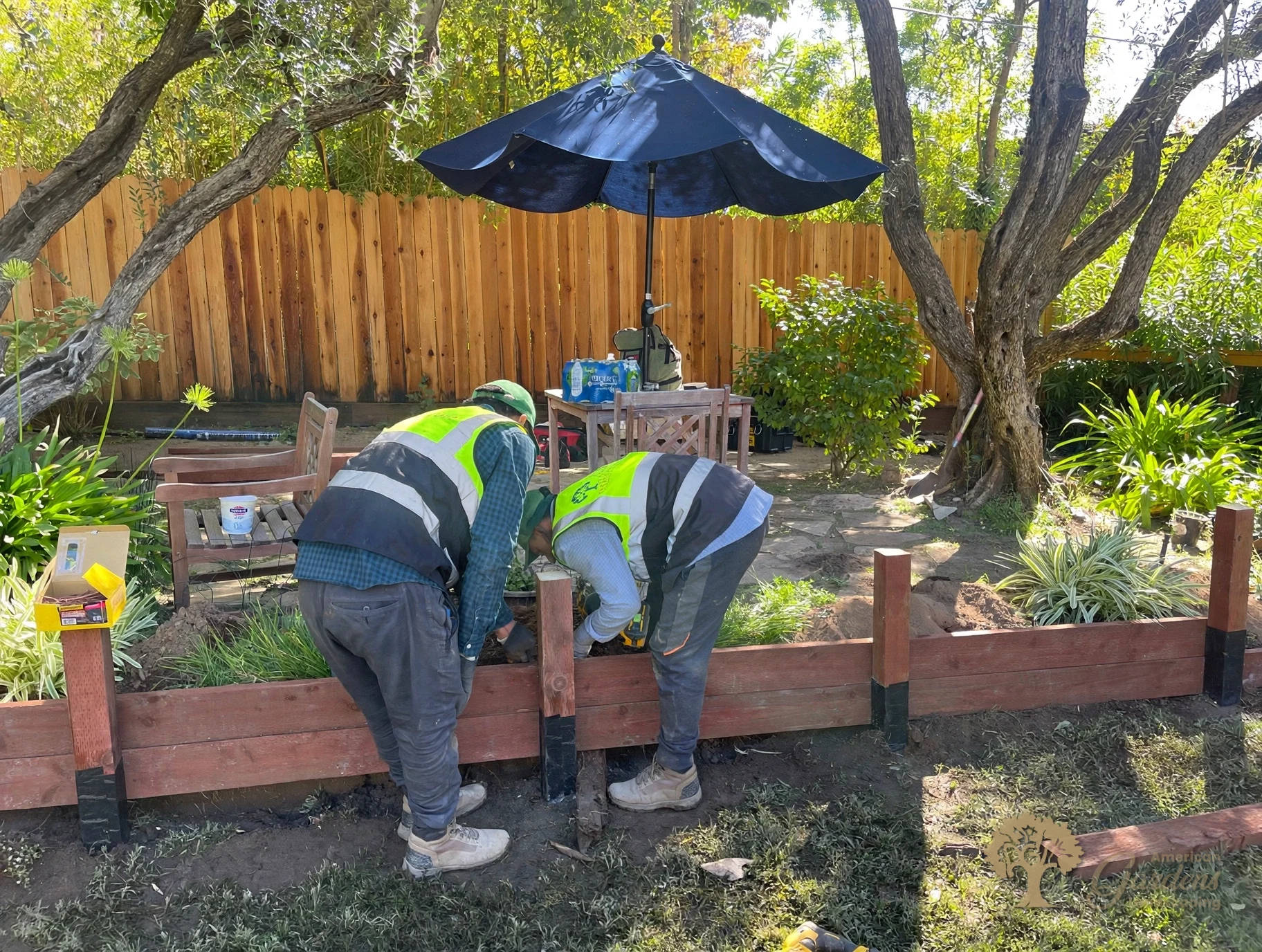Two American Gardens Landscaping crew members in company vests fastening pressure-treated timber boards onto posts wrapped in black waterproof wrap at the base, in Fair Oaks