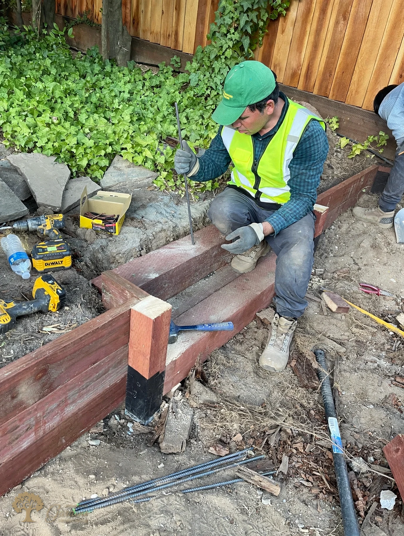 An American Gardens Landscaping crew member in a green company cap pinning the pressure-treated timber courses of a retaining wall with rebar in Fair Oaks
