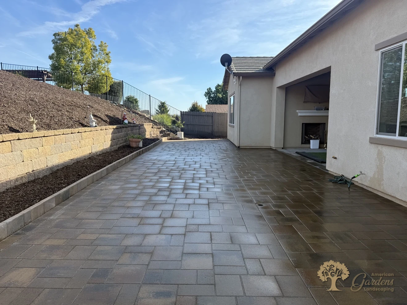 Freshly washed finished paver patio looking toward the covered patio with an outdoor fireplace, block retaining wall and bark hillside, in El Dorado Hills by American Gardens Landscaping
