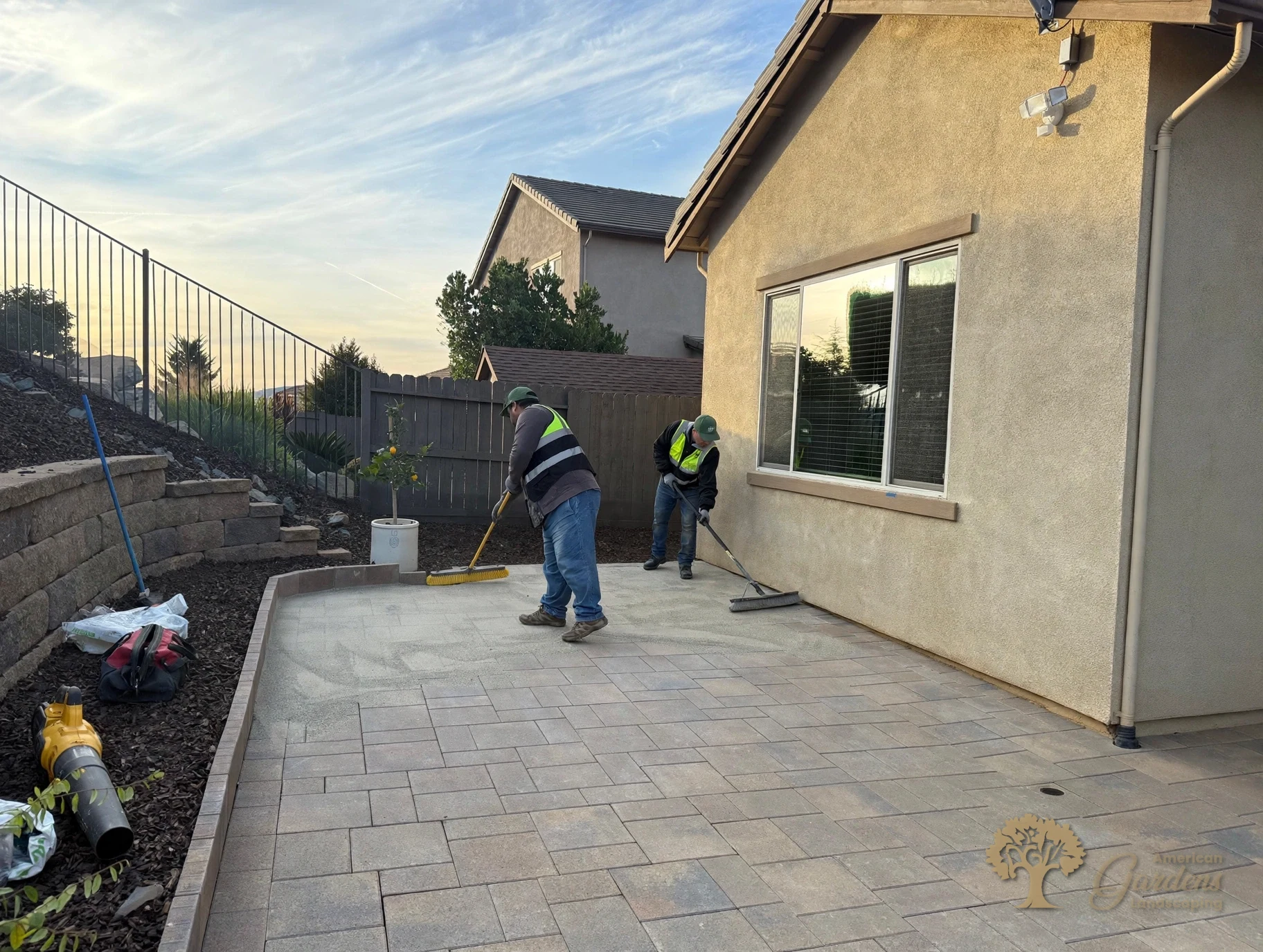 Two American Gardens Landscaping crew members in company hi-vis vests sweeping joint sand into a finished paver patio at sunset in El Dorado Hills