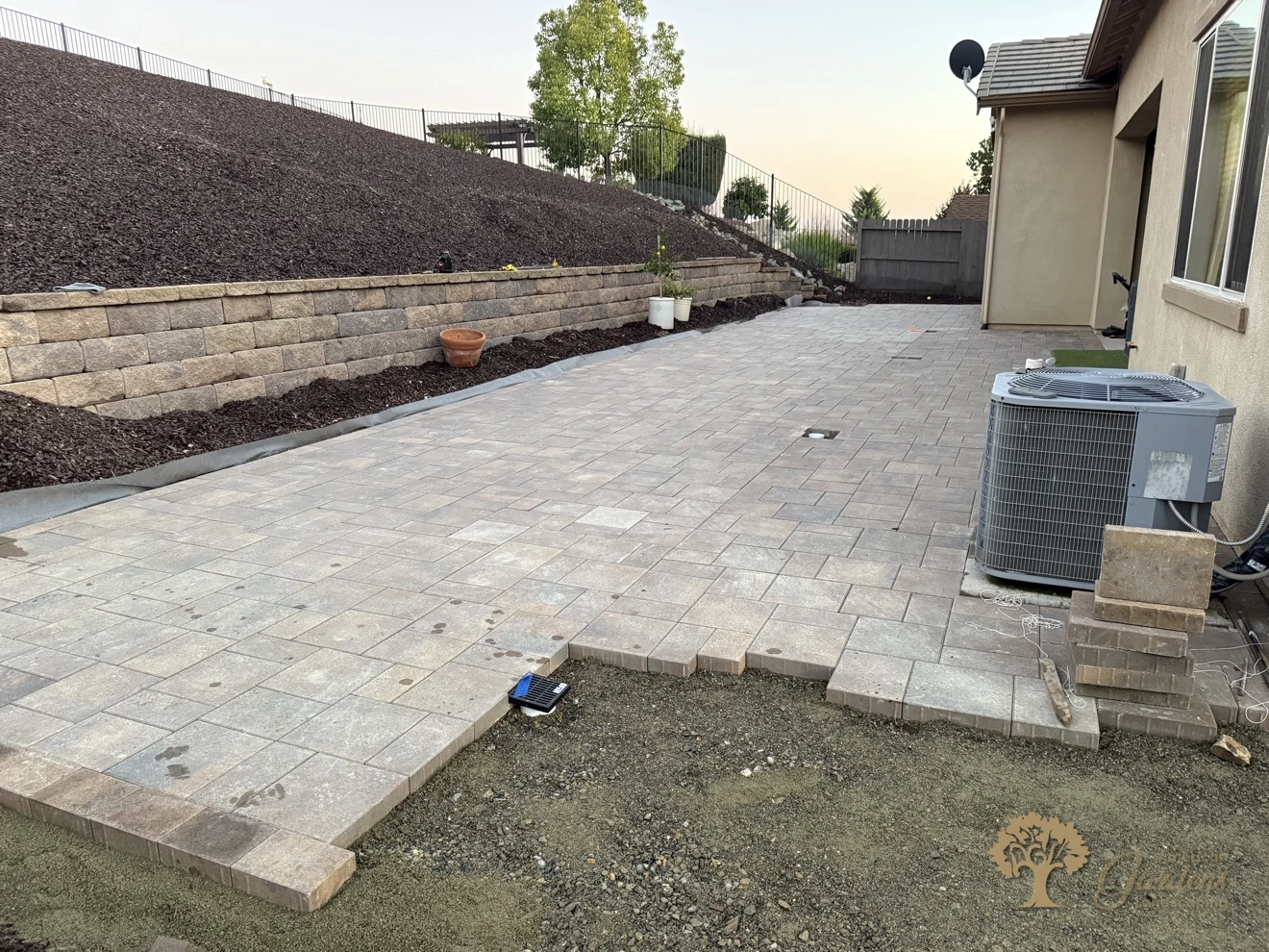 Long view of a finished paver patio at dusk beside a stucco house, with a block retaining wall and bark hillside, in El Dorado Hills by American Gardens Landscaping