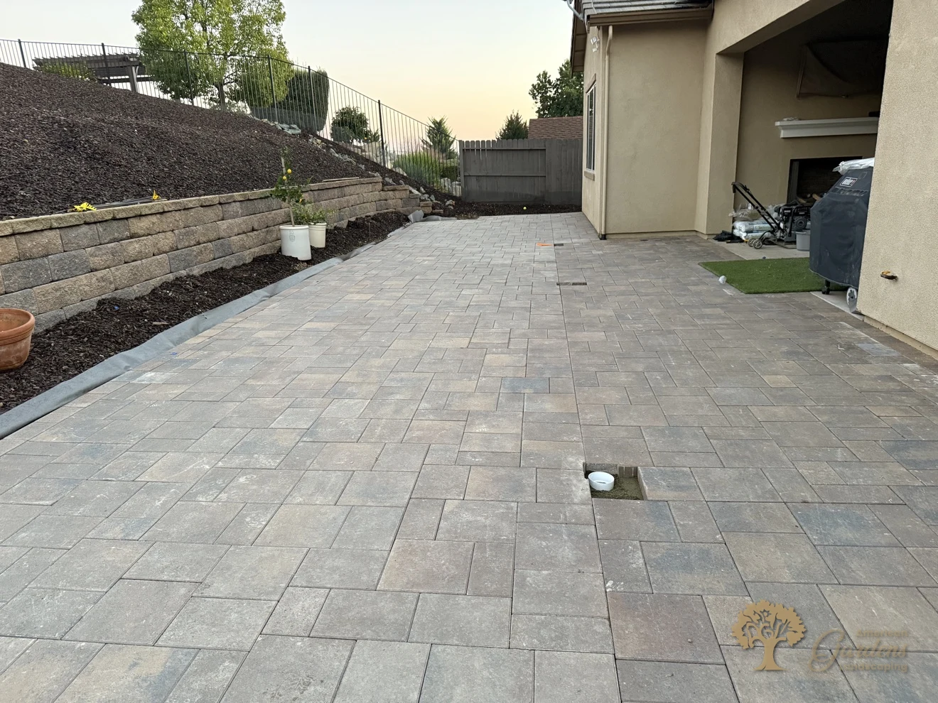 Finished paver patio at dusk looking toward the covered patio with an outdoor fireplace, block retaining wall and bark hillside, in El Dorado Hills by American Gardens Landscaping