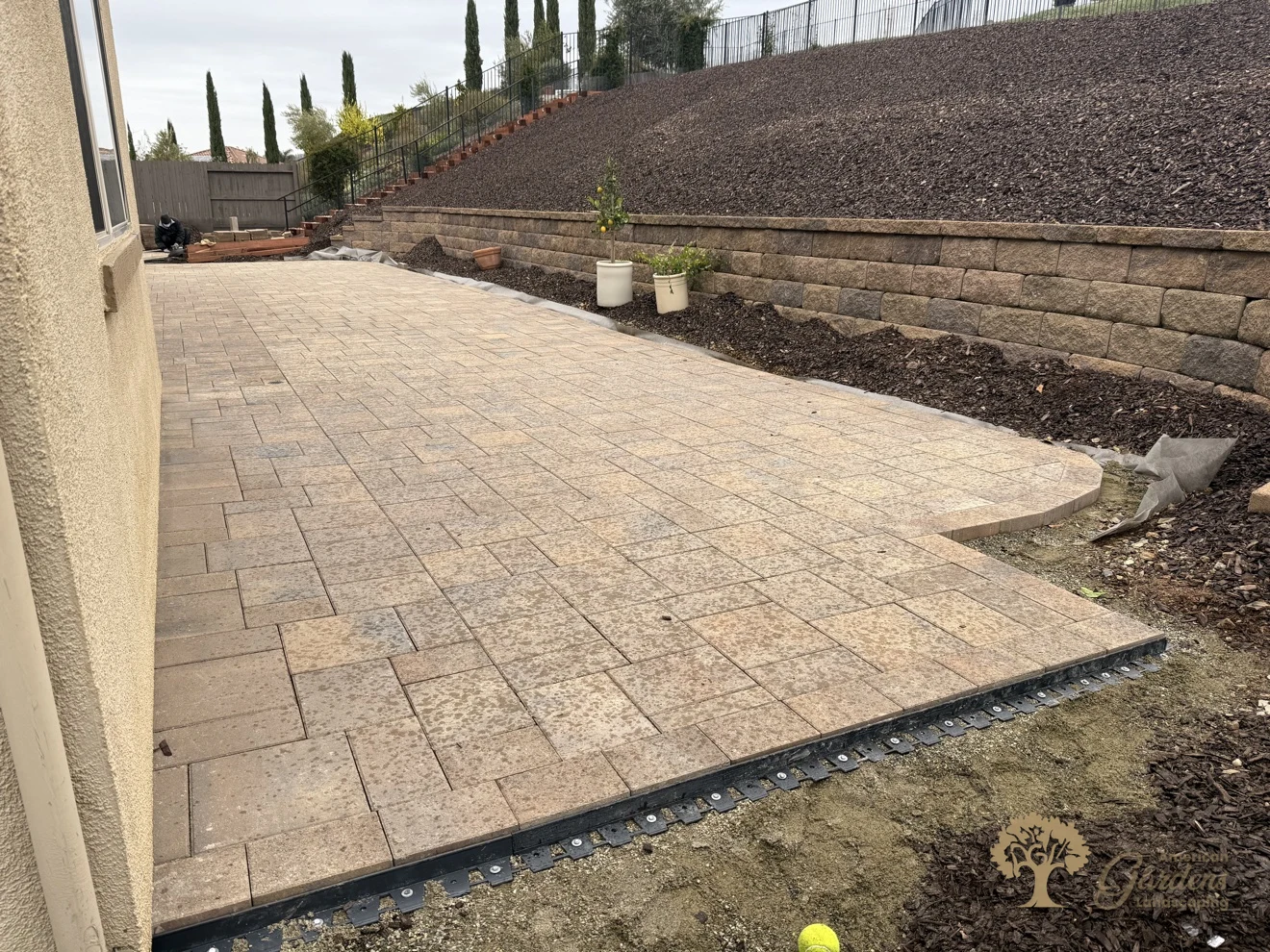 Finished paver patio in daylight showing its curved edge and black edge-restraint strip, with a block retaining wall and bark hillside, in El Dorado Hills by American Gardens Landscaping