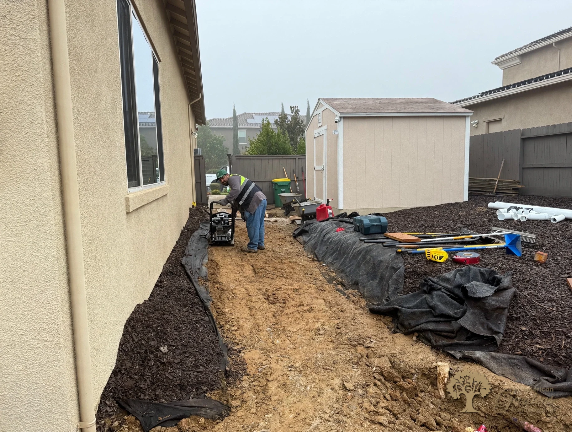 American Gardens Landscaping crew member in a company vest running a plate compactor over the base layer for a paver patio in El Dorado Hills