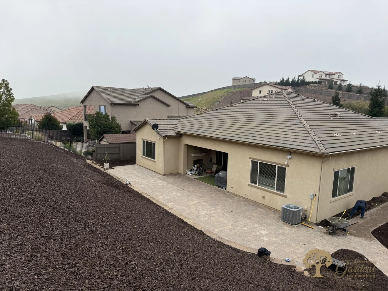 High-angle overview of a stucco house and its finished backyard paver patio with a bark hillside in El Dorado Hills by American Gardens Landscaping