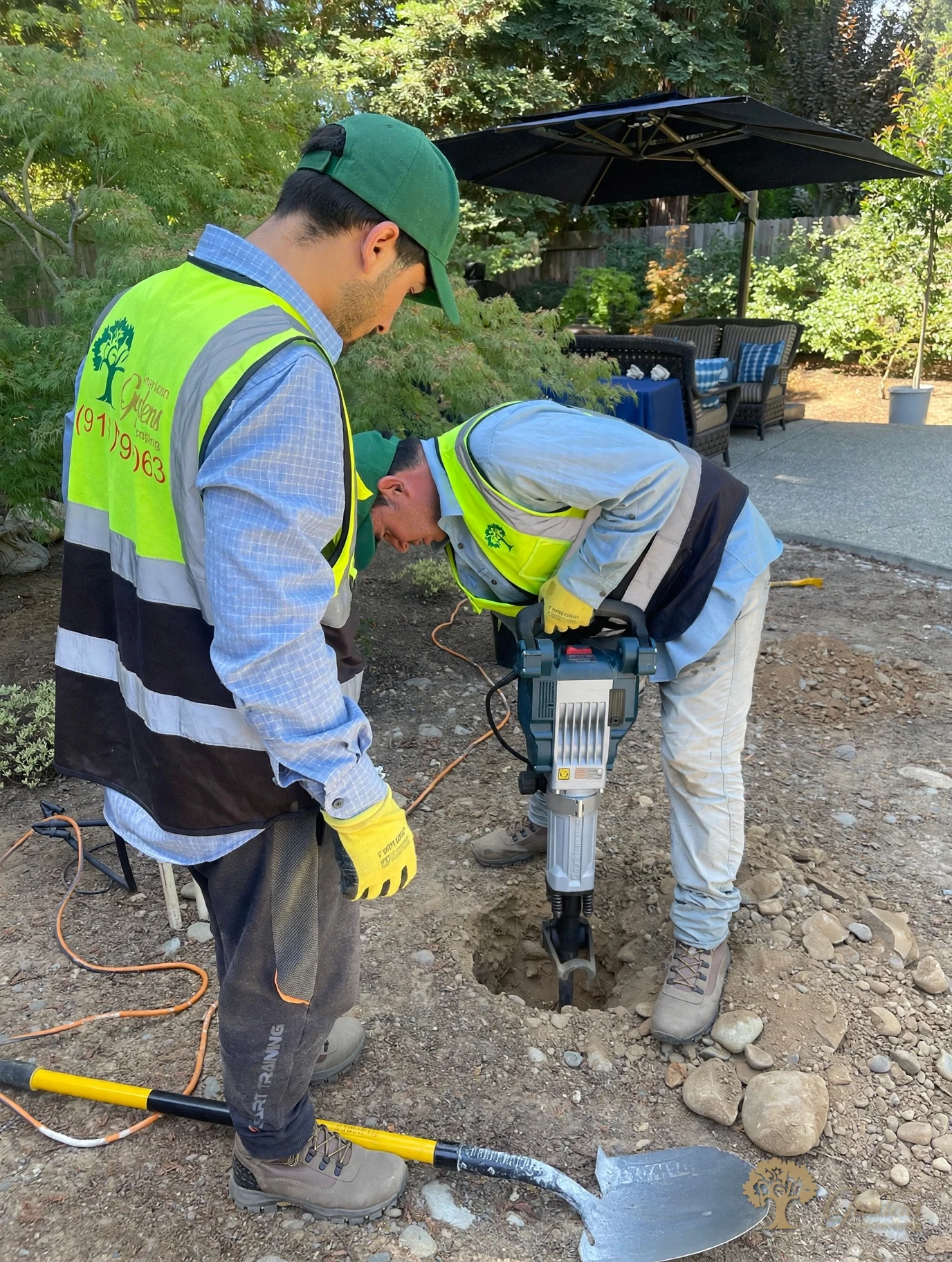 Two American Gardens Landscaping crew members in hi-vis vests and green caps jackhammering a planting hole in the rocky soil of a Gold River backyard