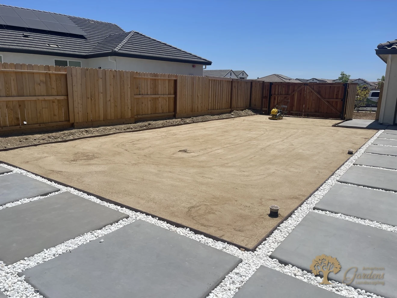 Wide view of a decomposed granite area being compacted smooth with a plate compactor, beside concrete pavers with white-rock joints, by American Gardens Landscaping