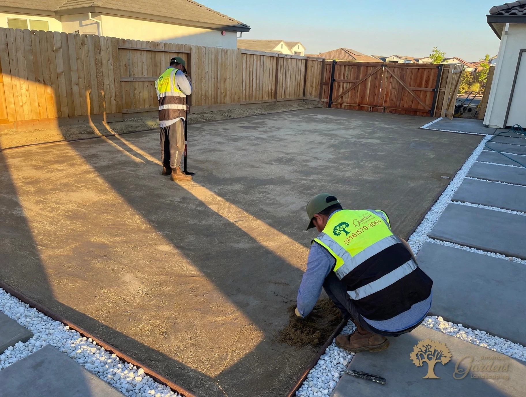 Two American Gardens Landscaping crew members in company vests hand-finishing the edges of a compacted decomposed granite pad at golden hour